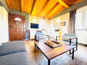 Ein gemütliches Wohnzimmer mit Holzbalkendecke, Fernseher, Schachbrett und Kamin.