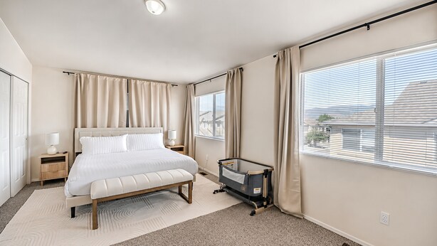 Quarto com cama, mesas de cabeceira, berço, janela com persianas e vista para edifícios.