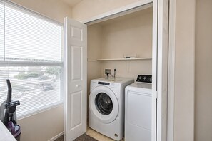A laundry room with a front-loading washer and dryer, a vacuum cleaner, and a window with blinds. - Family 3BR/3.5BA W/ Cinema Near USAFA + Garden of the Gods (Colorado Springs)