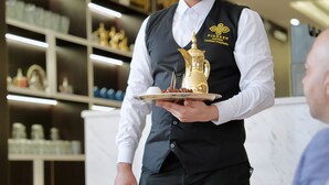 Um garçom segurando uma bandeja com um jarro dourado e comida, em um salão bem iluminado com prateleiras ao fundo.