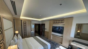 Quarto de hotel com cama, mesa de cabeceira, luminária, televisão e uma janela grande.