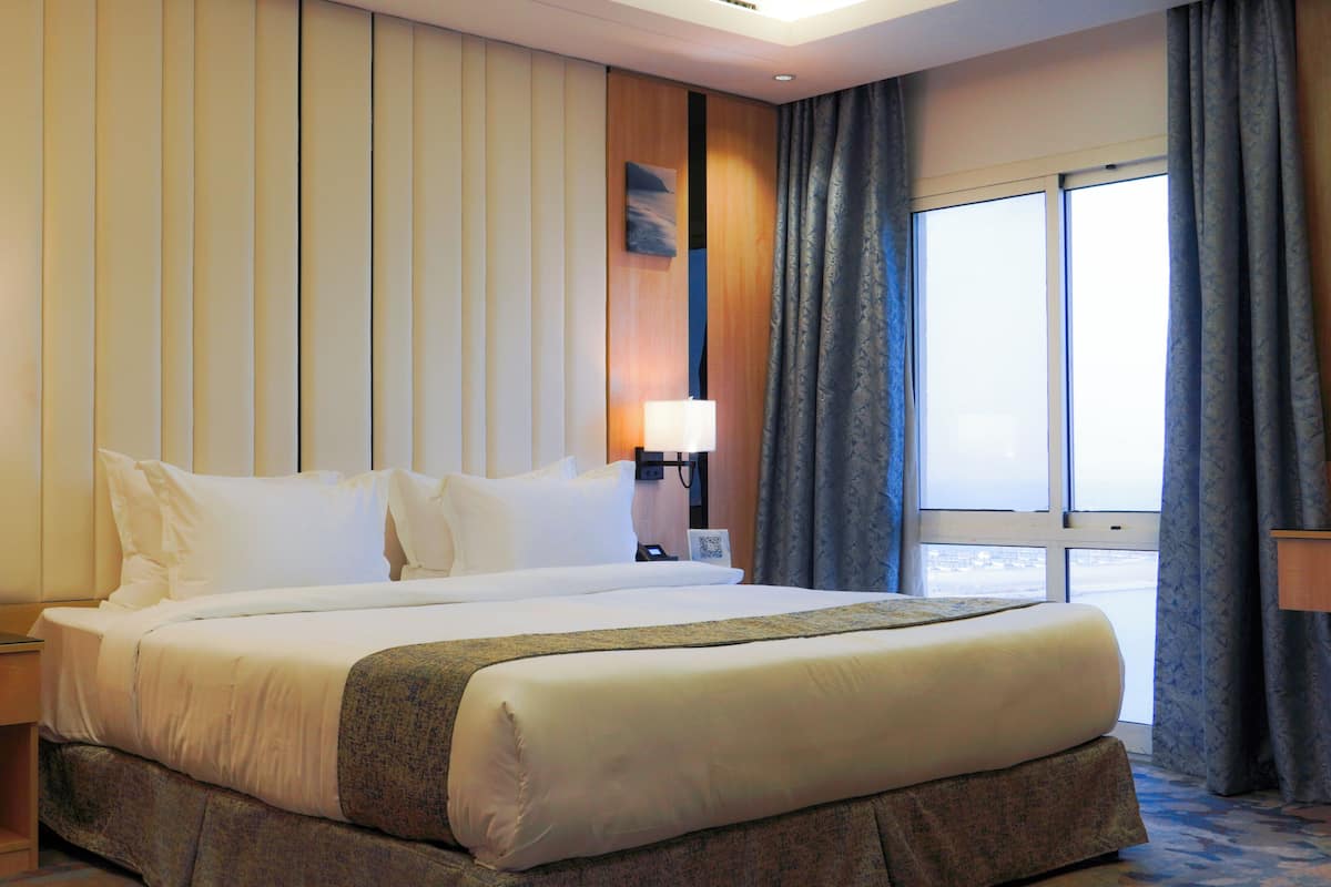 A hotel room with a large bed, two bedside tables, a nightstand, a window with curtains, and a wall with vertical panels.
