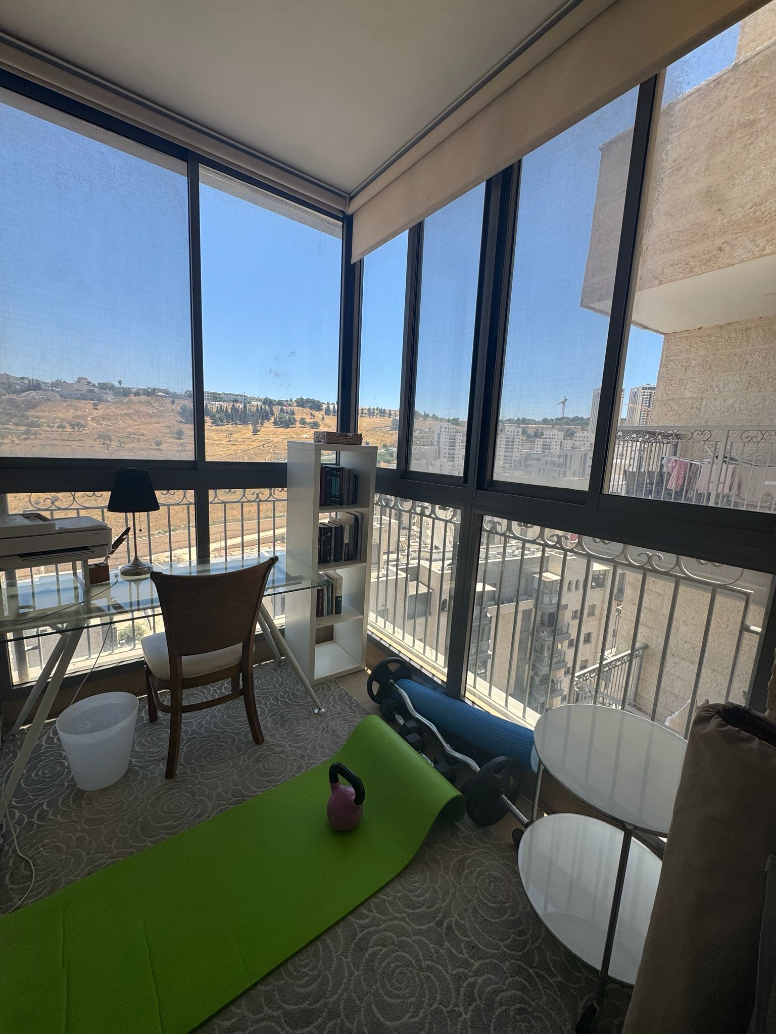Ein Balkon mit Blick auf eine hügelige Landschaft, mit einem Tisch, einem Stuhl, einem Bücherregal und einer Kettlebell auf einer Matte.