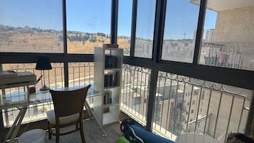 Ein Balkon mit Blick auf eine hügelige Landschaft, mit einem Tisch, einem Stuhl, einem Bücherregal und einer Kettlebell auf einer Matte.