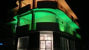 En bygning med flere etager og en buet facade, oplyst af grønne og gule lys, samt en tydelig indgang med glasdøre.