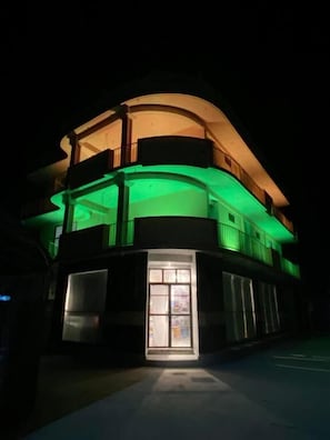 A multi-story building with a curved facade, illuminated by green and yellow lights, and a visible entrance with glass doors. - Bonthi Grand (Gemanafushi)