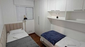 Una habitación pequeña y limpia con dos camas, un escritorio, un ventilador de techo y paredes blancas.