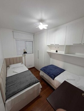 Ein kleines, sauberes Zimmer mit zwei Betten, einem Schreibtisch, einem Deckenventilator und weißen Wänden.