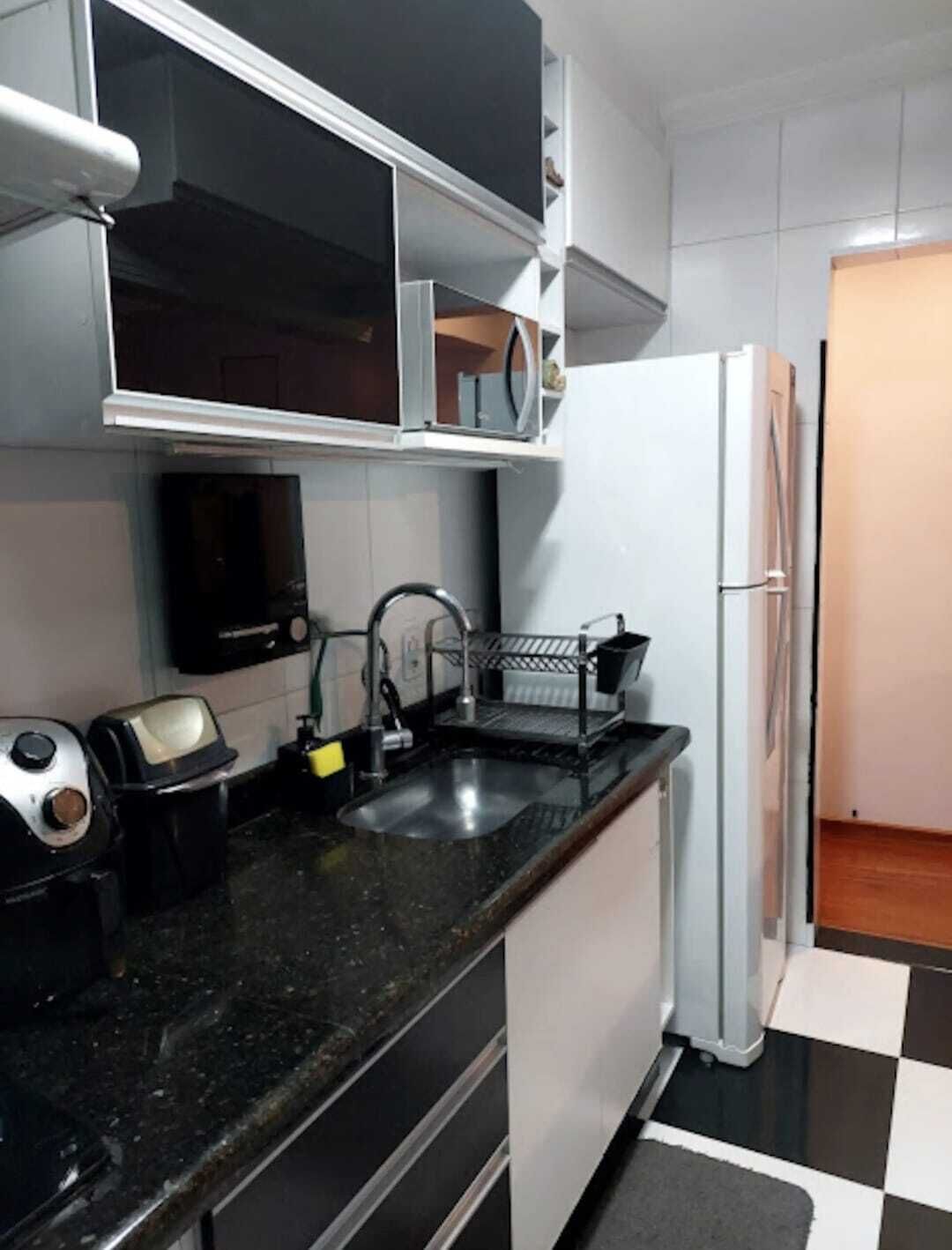 Cozinha compacta com bancada preta, armários brancos, micro-ondas embutido e geladeira.