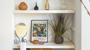 Une étagère blanche ornée d’une œuvre d’art aux couleurs jaune et noir, d’un vase avec un tournesol, d’un tableau encadré, d’un petit miroir, d’une plante en pot et d’un panier tressé.