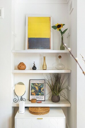 Ein weißes Regal mit einer gelb-schwarzen Grafik, einer Sonnenblumenvase, einem gerahmten Bild, einem kleinen Spiegel, einer Topfpflanze und einem gewebten Korb.