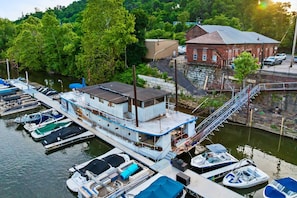Une péniche amarrée, dotée d’une terrasse couverte, d’un escalier menant à l’étage supérieur et d’un quai où sont accostés plusieurs petits bateaux.