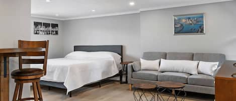 Una habitación con una cama, un sofá gris, una silla de madera, una mesita redonda y un cuadro en la pared.
