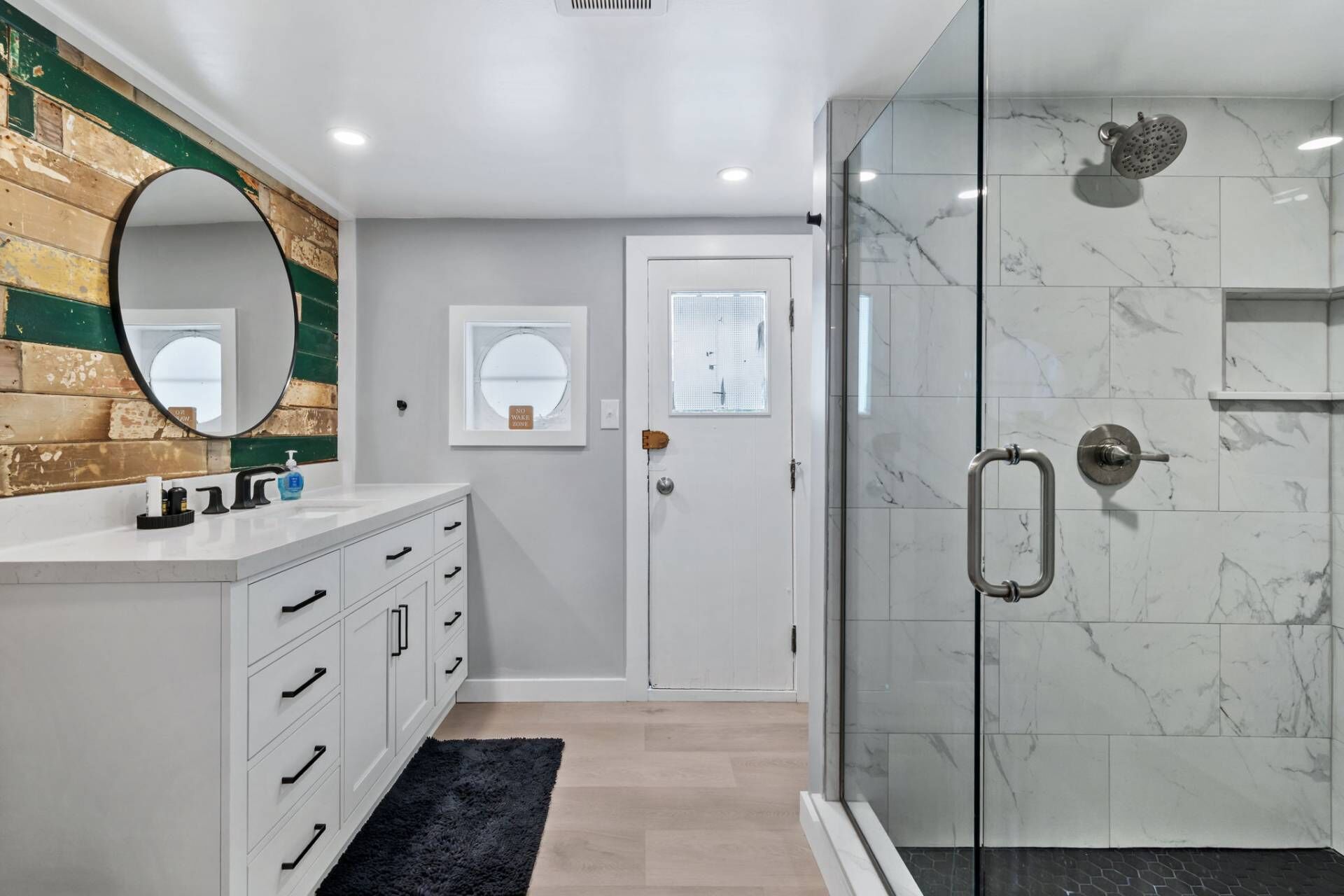 Une salle de bain moderne avec une douche à l’italienne en verre, un meuble-lavabo blanc avec des tiroirs, un miroir rond et un mur d’accent en bois.