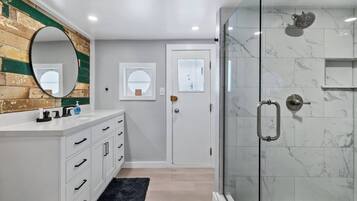 Une salle de bain moderne avec une douche à l’italienne en verre, un meuble-lavabo blanc avec des tiroirs, un miroir rond et un mur d’accent en bois.