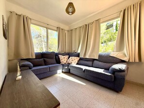 Ein Wohnzimmer mit einer Eckcouch, einem Holztisch und großen Fenstern mit Vorhängen.