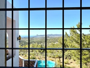 Une vue à travers une fenêtre à carreaux, donnant sur un paysage composé d’arbres, d’une piscine et de collines lointaines.