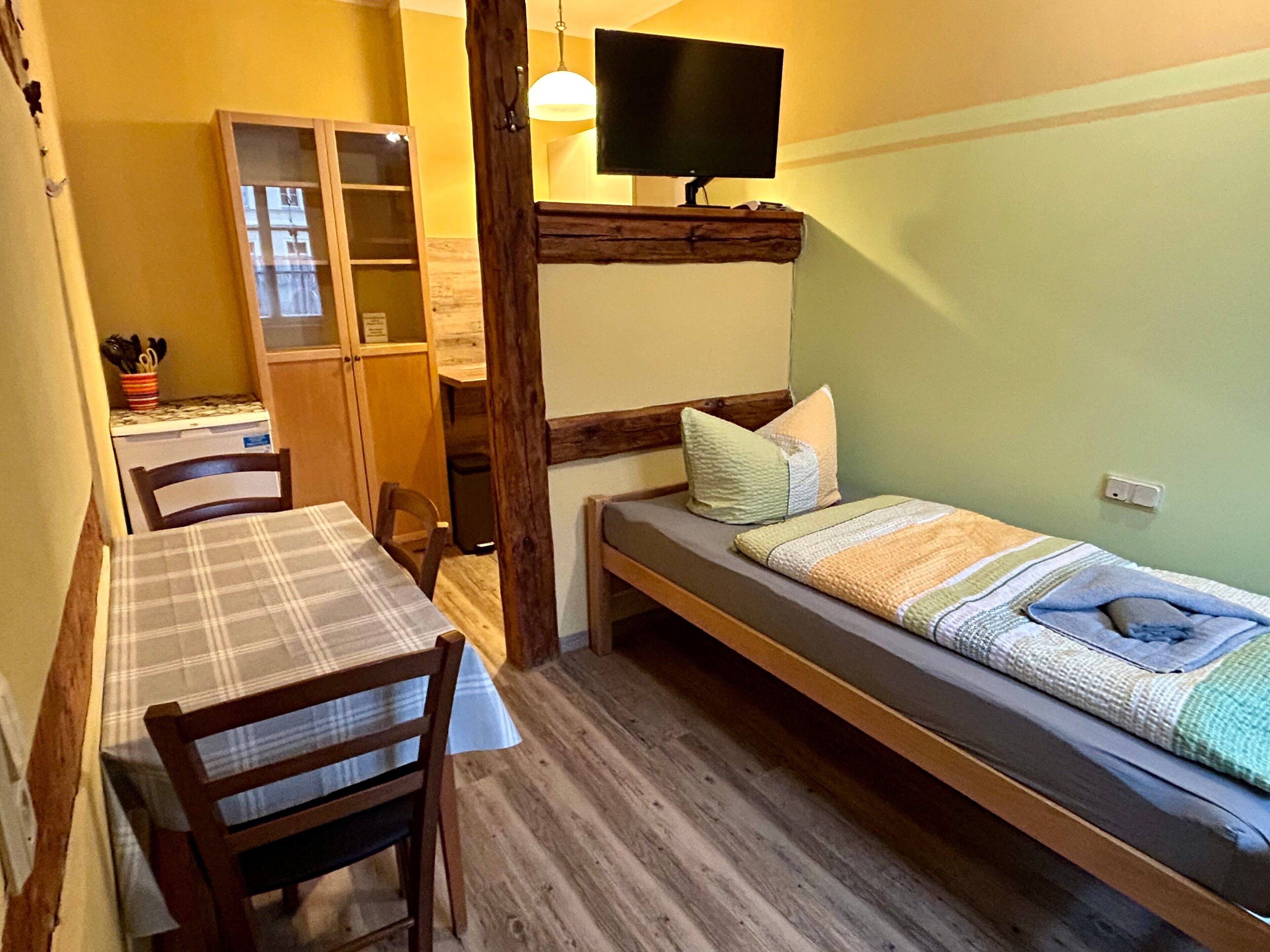 Een kleine kamer met een bed, een tafel en een stoel. Er is een televisie aan de muur bevestigd.