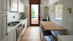 A compact kitchen with a stove, oven, and washing machine, a wooden table, and a view through a door to an outdoor area. - Stylish Lido Beach Apartment with Terrace, Shared Garden & Steps to the Sea (Lido)