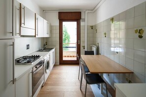 Eine kleine Küche mit Herd, Backofen und Waschmaschine, einem Holztisch und Blick durch eine Tür in einen Außenbereich.