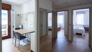A hotel room with a wooden floor, a bed with a white bedspread, a small table with chairs, and a window with curtains. - Stylish Lido Beach Apartment with Terrace, Shared Garden & Steps to the Sea (Lido)