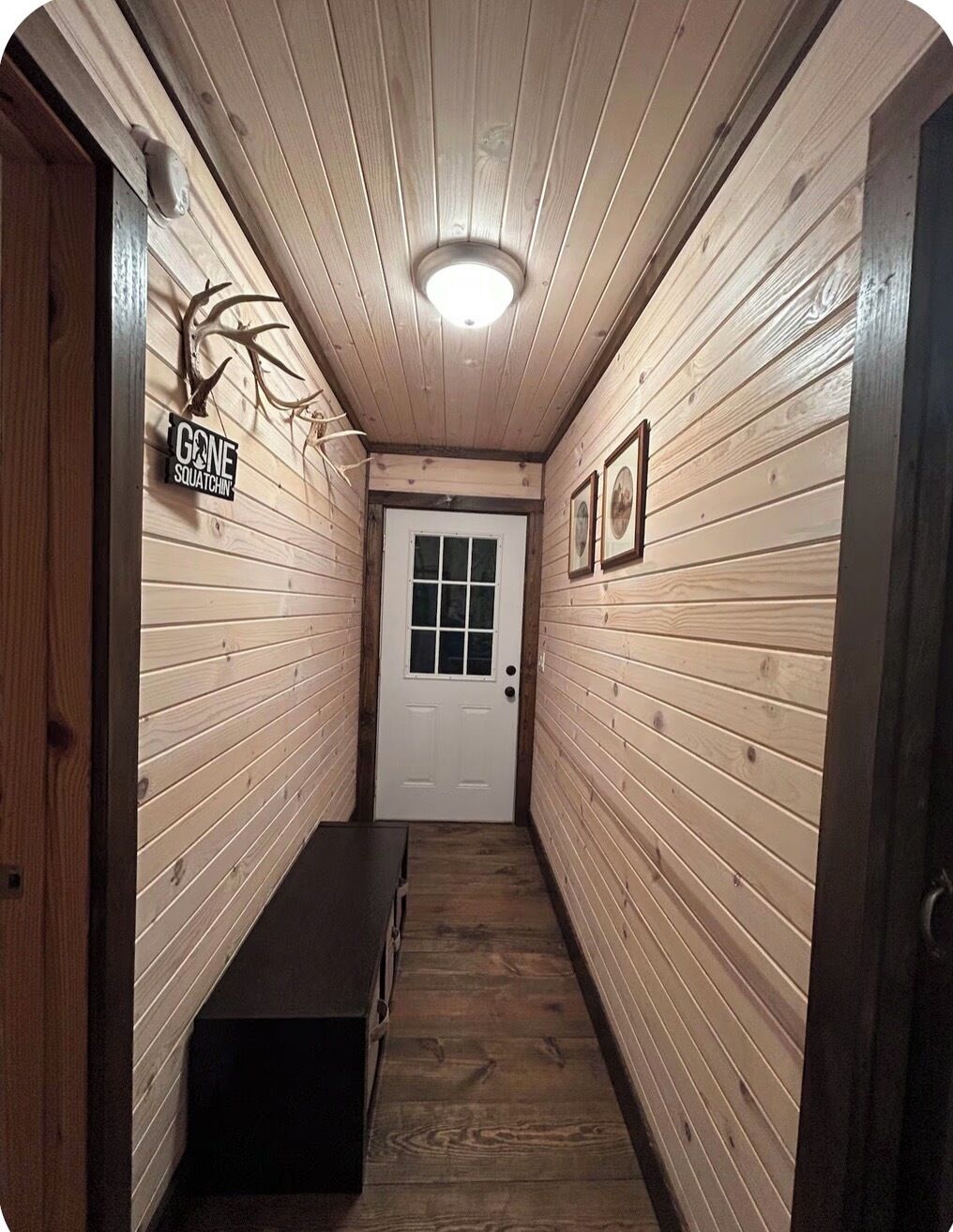 A narrow hallway with wooden walls, a bench, a door at the end, and a sign on the left wall.