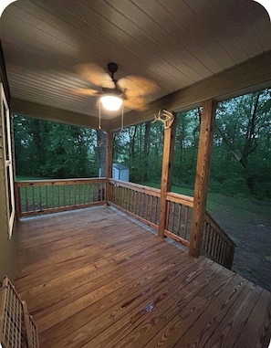 A wooden deck with a railing, a ceiling fan, and a view of a wooded area.