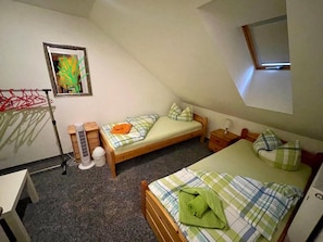 Eine kleine, einfache Unterkunft mit schrägen Wänden, einem Bett mit grün-weiß karierter Decke, einem Nachttisch mit Lampe und einem Fenster, das natürliches Licht hereinlässt.