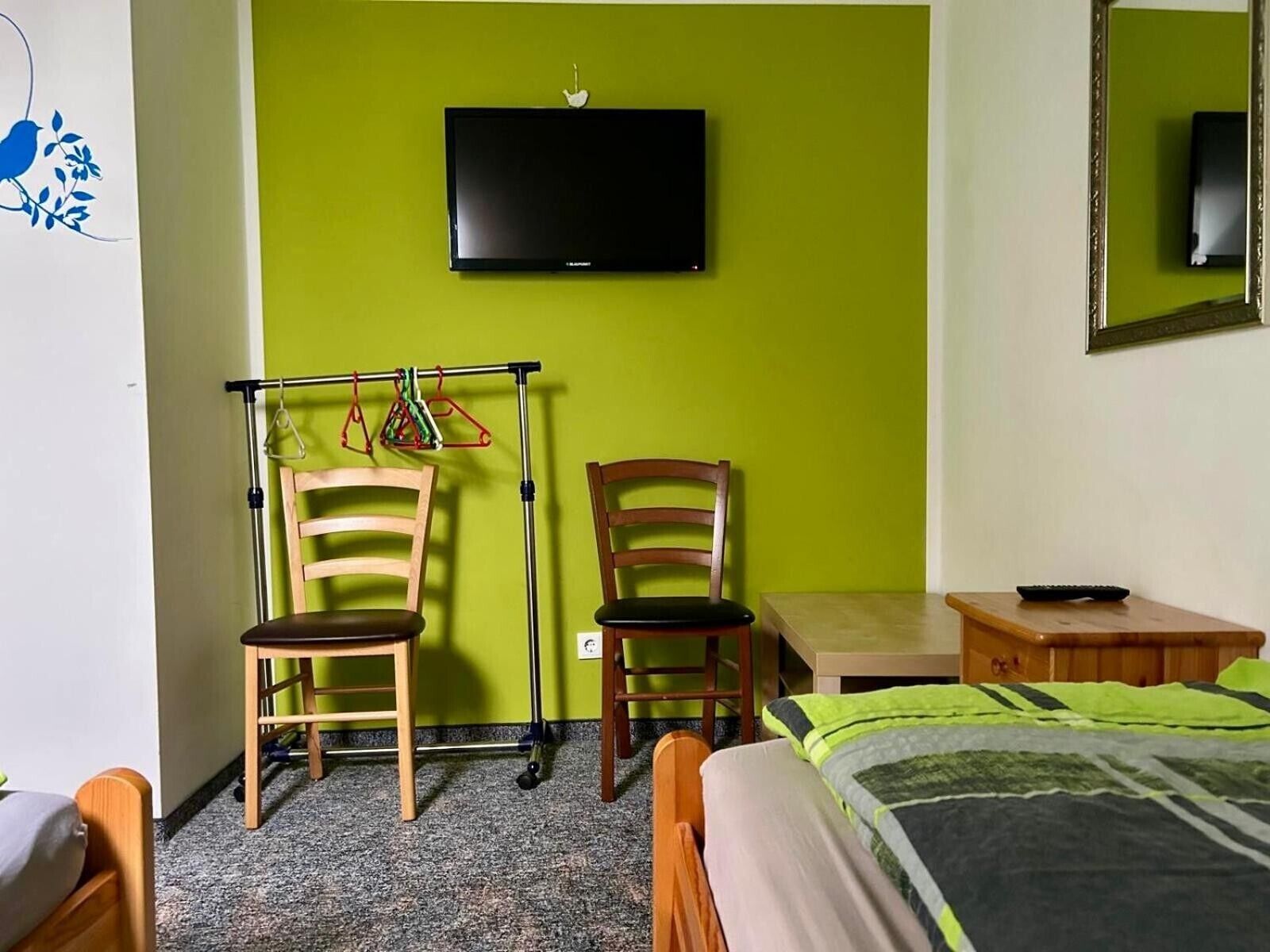 Een kamer met een bed, twee houten stoelen, een kleine tafel, een televisie bevestigd aan een groene muur en een spiegel aan de rechterkant.