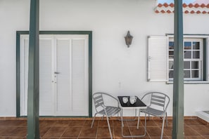 Eine Terrasse mit zwei Metallstühlen, einem kleinen Tisch, einer weißen Tür und einem Fenster mit Rollläden.
