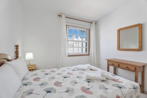 Ein Schlafzimmer mit Bett, Nachttisch mit Lampe, Holzkommode, Fenster mit Vorhängen und einem Spiegel an der Wand.