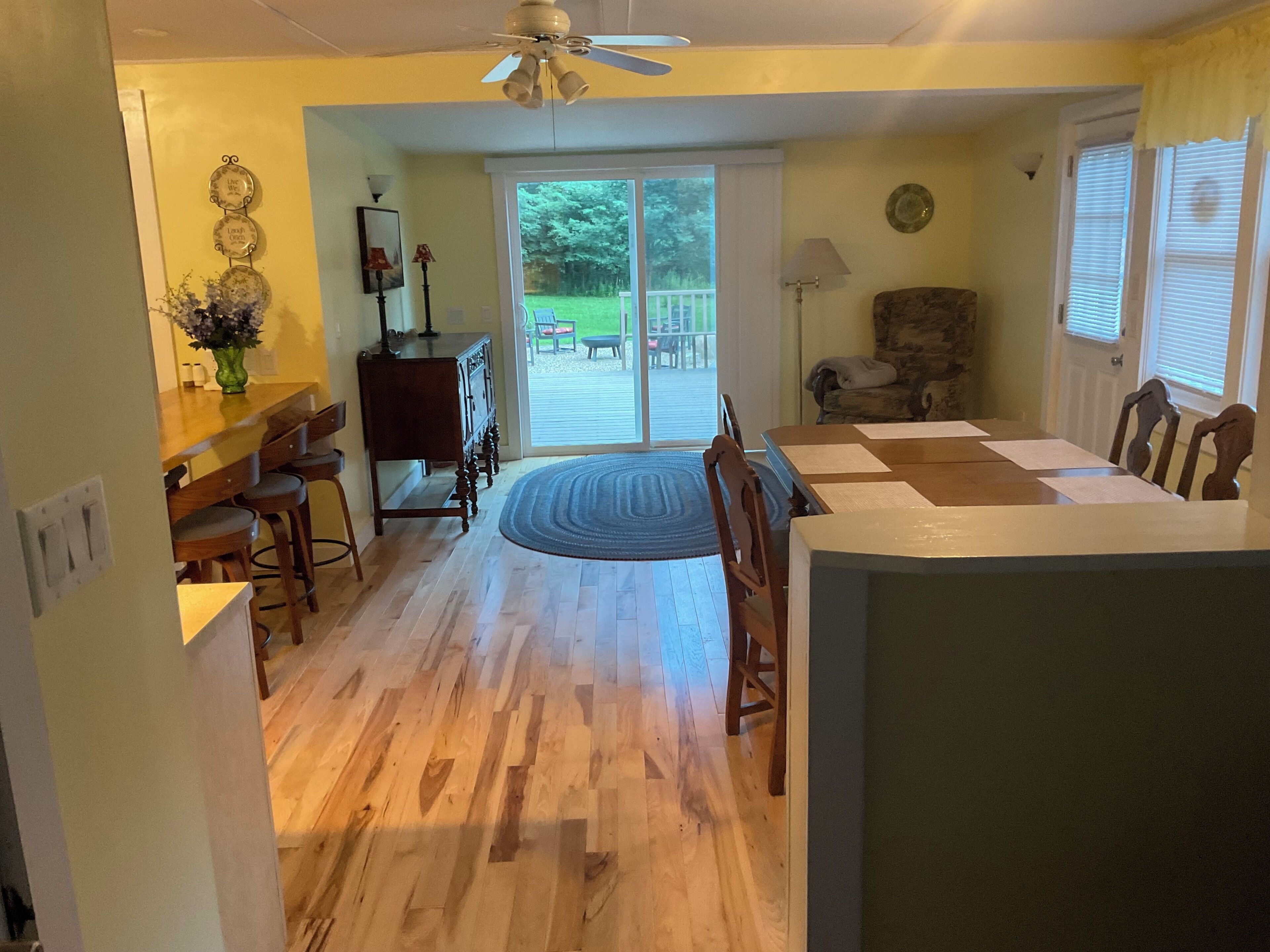 A dining area with a wooden table and chairs, a ceiling fan, and a view of the outdoors through a sliding glass door.