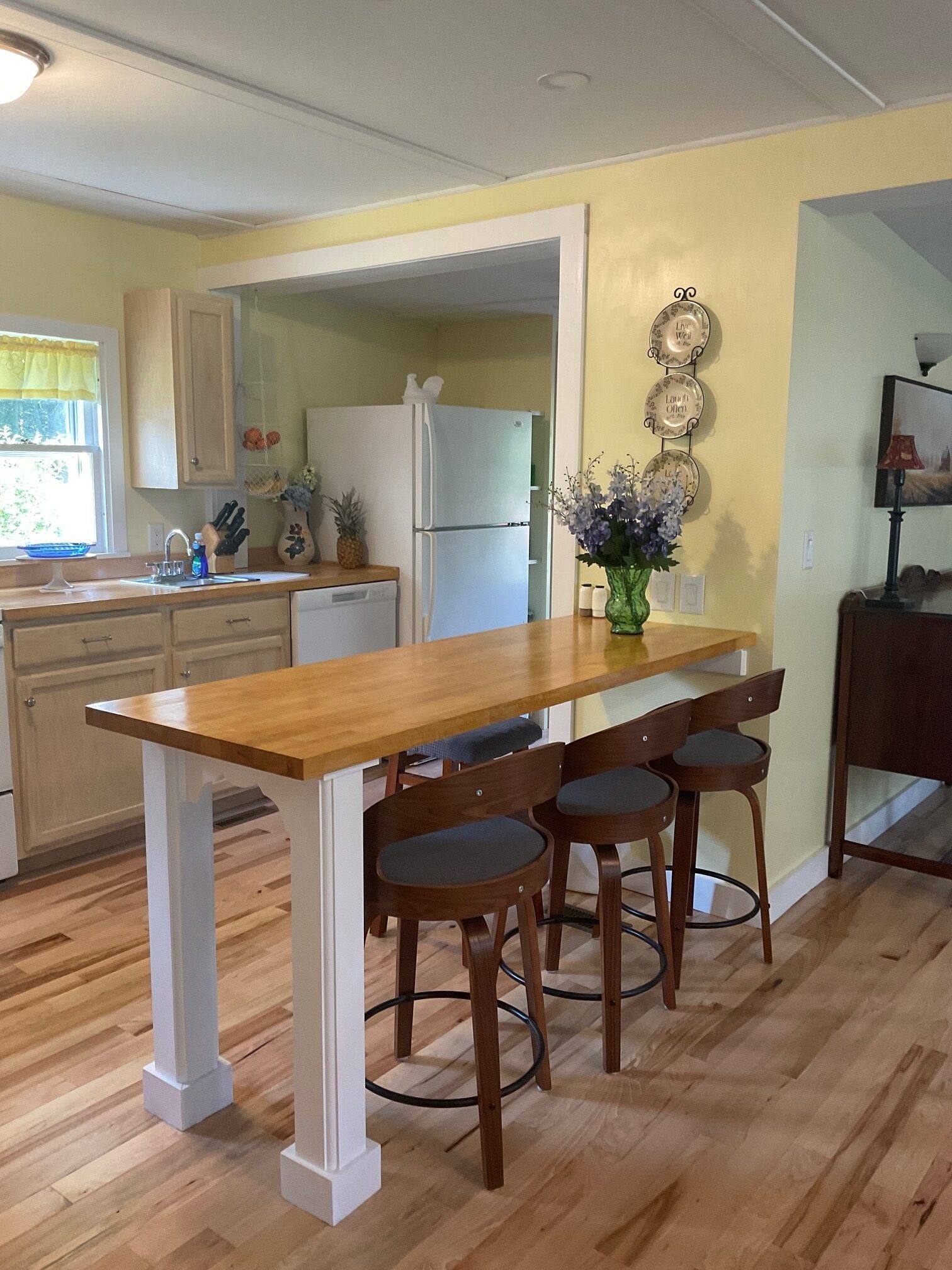 A kitchen with a wooden table and bar stools, a refrigerator, and a vase with flowers on the counter.