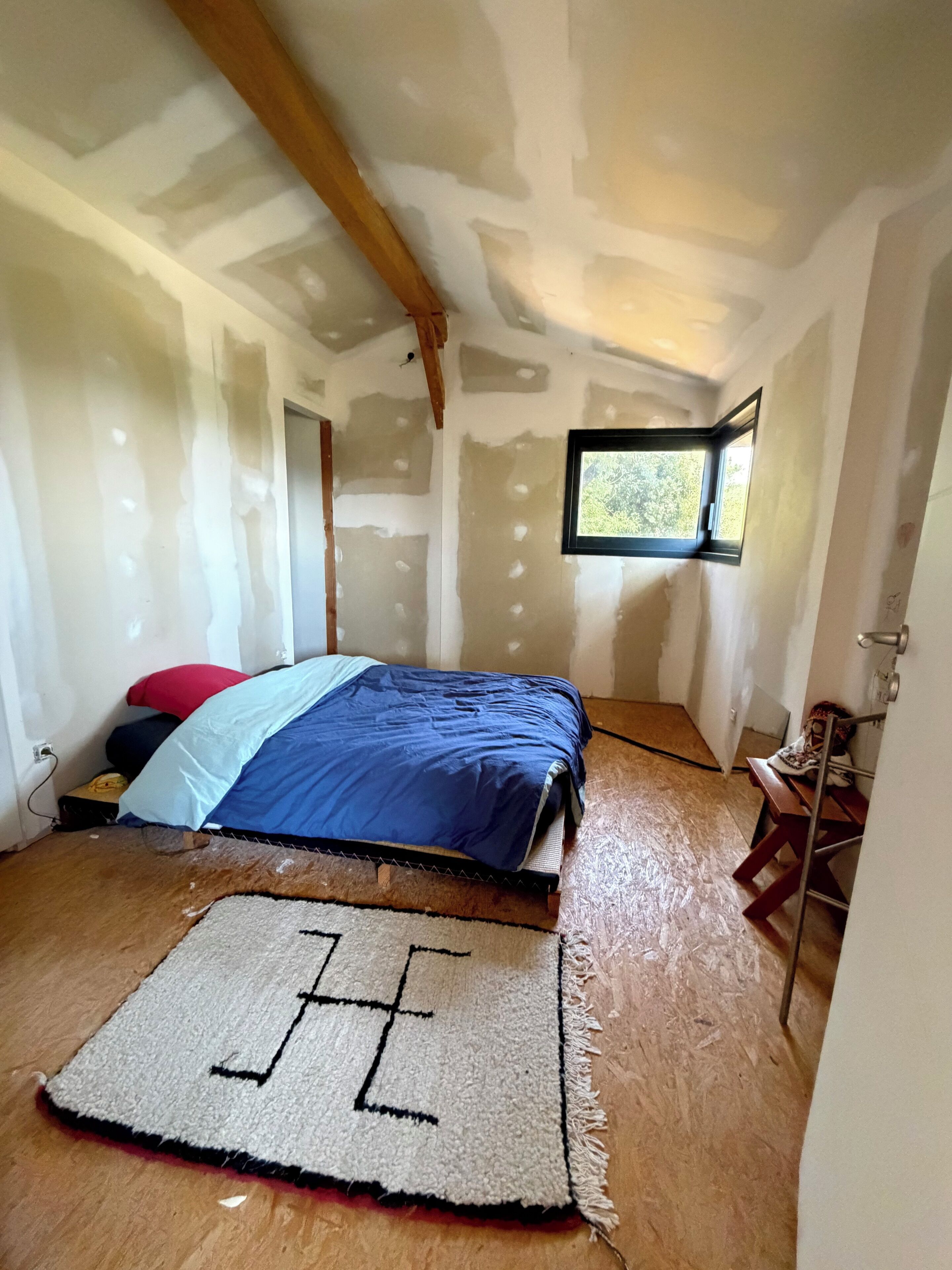 A room with exposed ceiling beams, a bed with a blue bedspread, a small window, and a wooden floor.