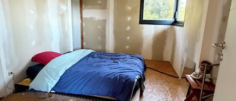 A room with exposed ceiling beams, a bed with a blue bedspread, a small window, and a wooden floor.