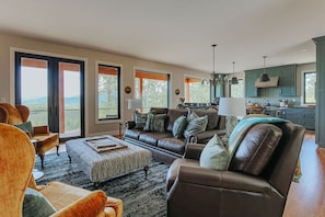 Ein geräumiges Wohnzimmer mit einer großen Couch, Hocker und Blick ins Freie durch große Fenster.