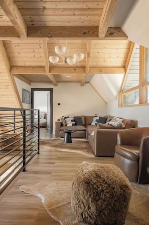 A loft-style living room with wooden beams, a leather sofa, a fur rug, and a metal railing. - Two Birds - expansive lake views and walk to lake (Lake Arrowhead)