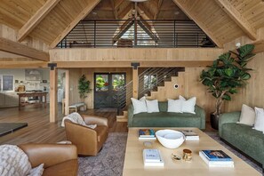A spacious loft with a high ceiling, wooden beams, a green sofa, a coffee table, and a staircase leading to an upper level. - Two Birds - expansive lake views and walk to lake (Lake Arrowhead)