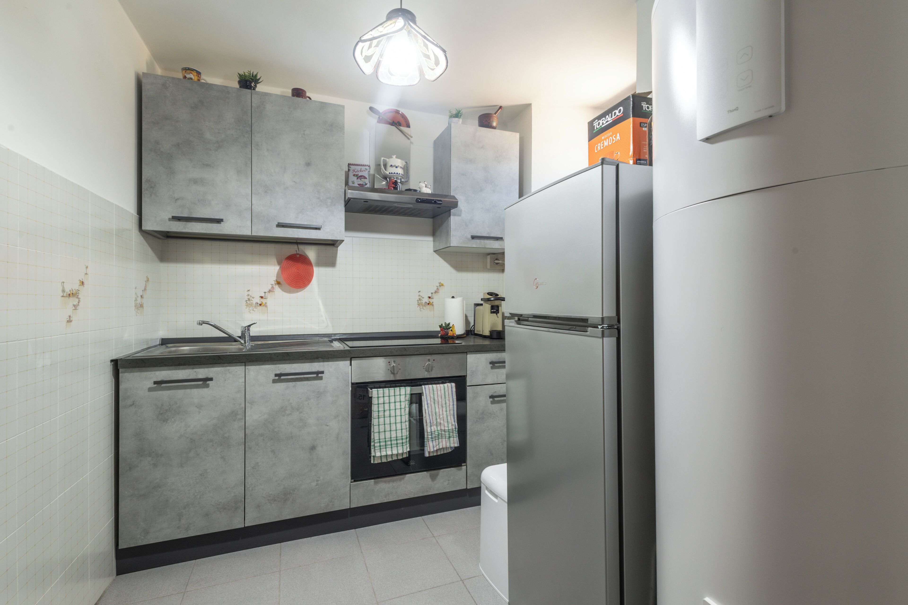 A kitchen with grey cabinets, a stainless steel refrigerator, a built-in oven, and a hanging light fixture.