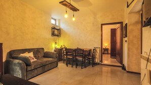 Una sala de estar con sofá, mesa de comedor y sillas, una escalera que conduce a otra habitación, y una lámpara fijada a la pared.