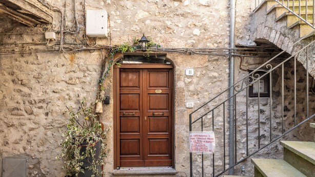 Edificio de piedra con puerta de madera, una escalera de metal y un cartel con texto.