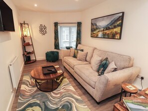 Ein Wohnzimmer mit einer Couch, einem Couchtisch, einem Bücherregal, einem Fenster mit Vorhängen und einem Bild an der Wand.