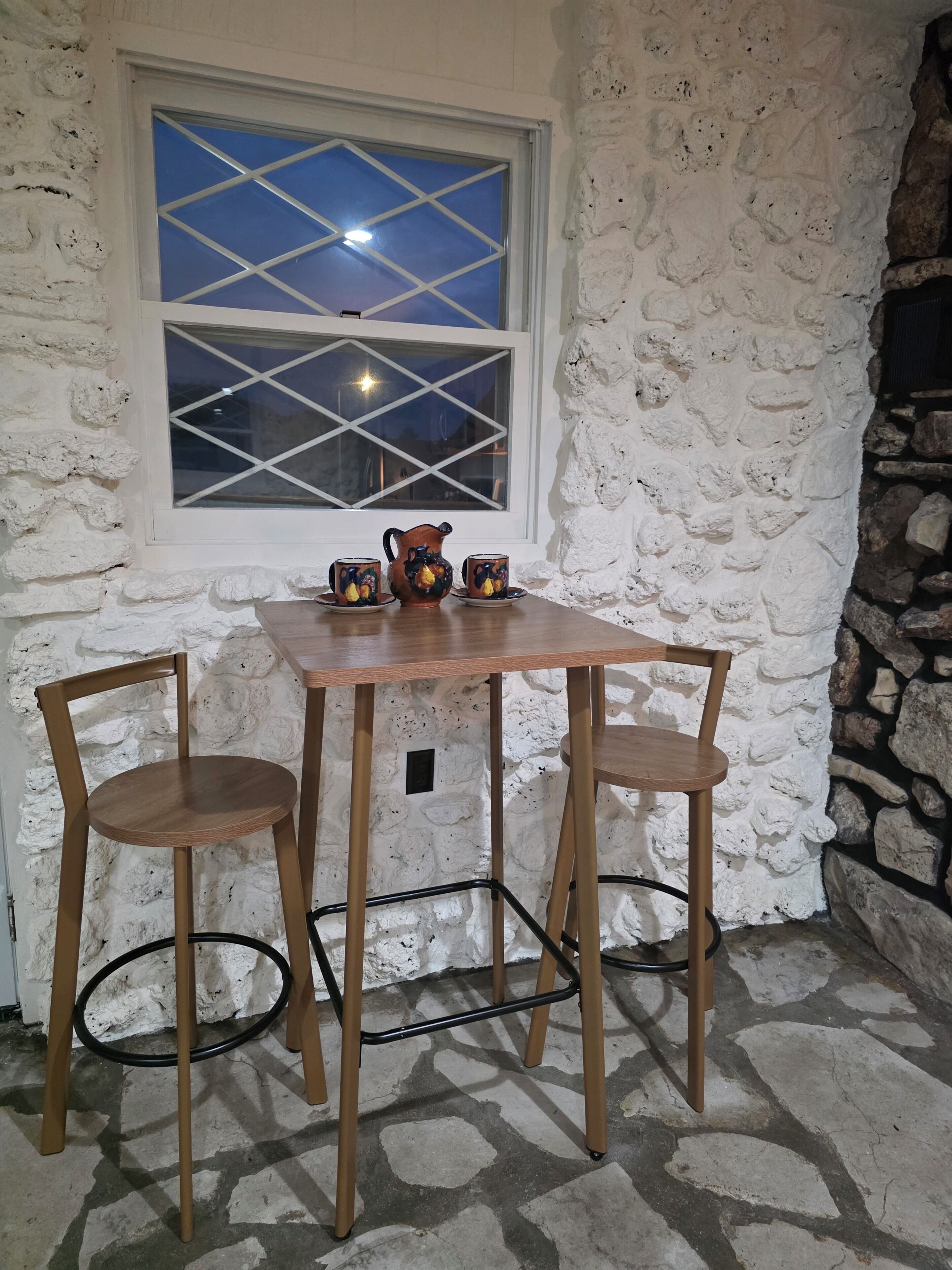 A stone-walled room with a table and two chairs, a window with a view of the night sky, and a decorative pitcher.