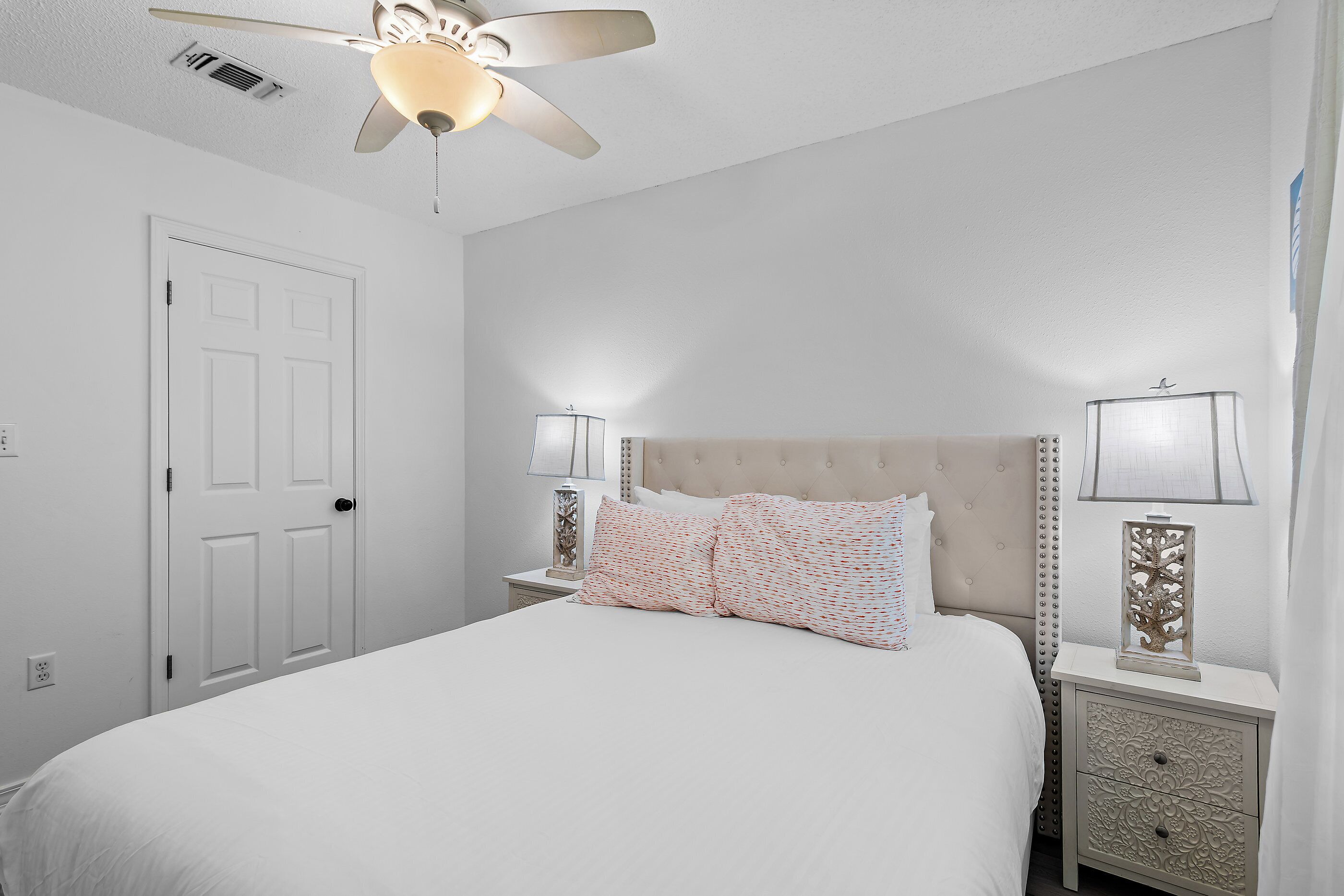 A bedroom with a bed, nightstands, lamps, a ceiling fan, and a door.
