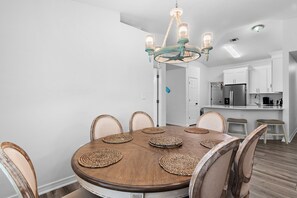 Un comedor con una mesa redonda de madera, seis sillas, una araña y una cocina al fondo.