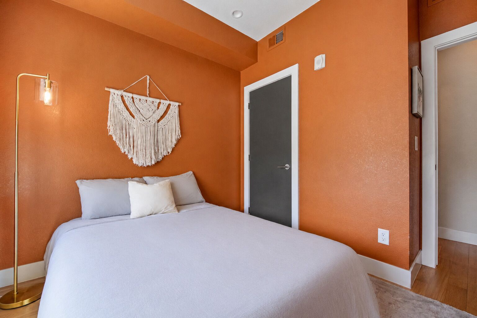 Ein Schlafzimmer mit einem Bett, einer Wanddekoration aus Makramee, einer Stehlampe und einer Tür.