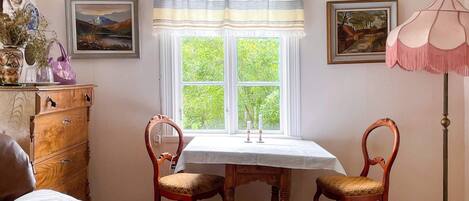A room with a wooden dresser, two chairs, a table, a window with a view of greenery, and a floor lamp.