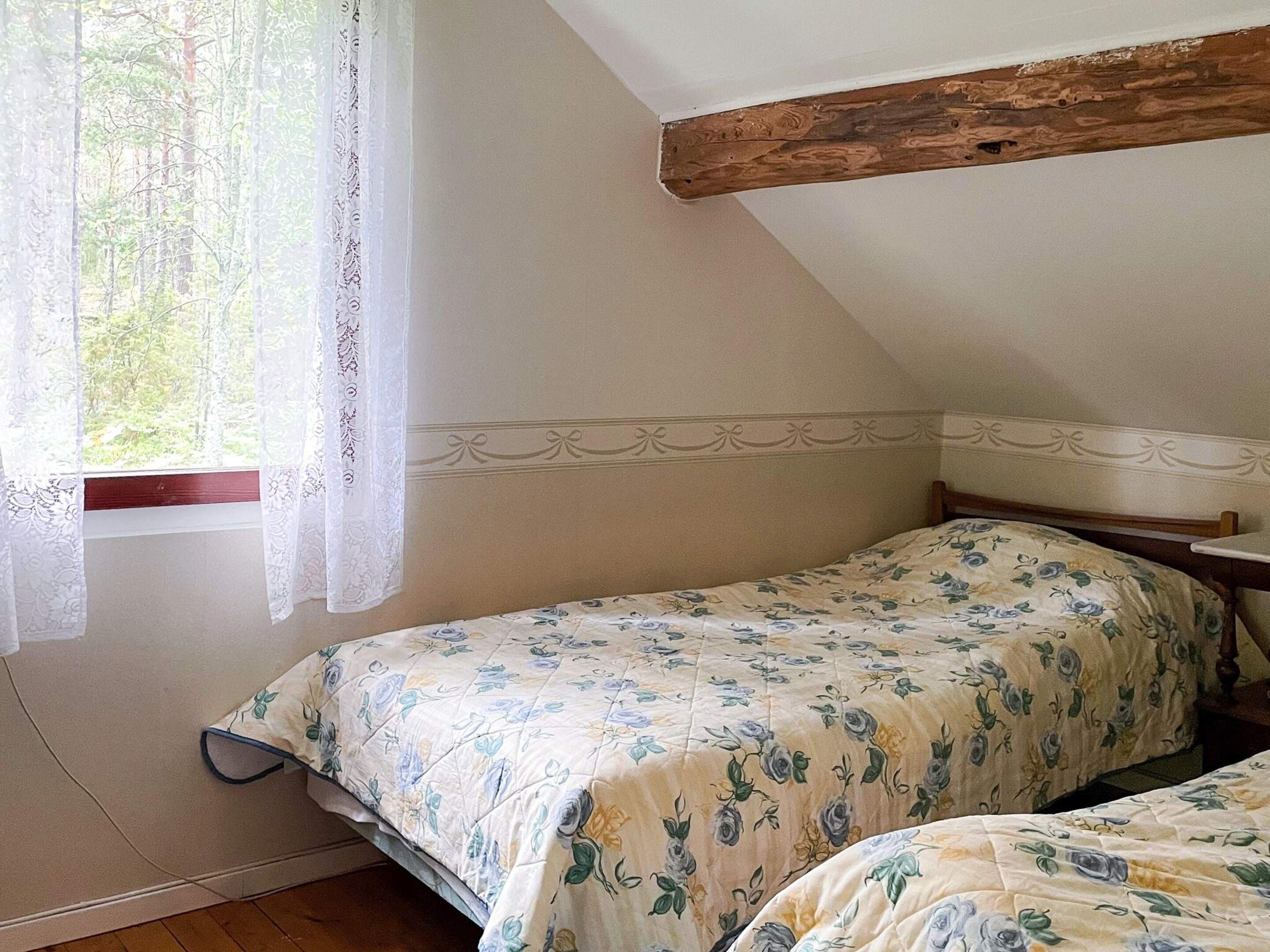 A bedroom with two single beds, a wooden ceiling beam, a window with lace curtains, and a floral bedspread.