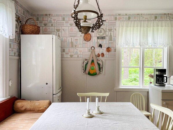 A kitchen with a dining table set for two, a refrigerator, and a window with a view of greenery.