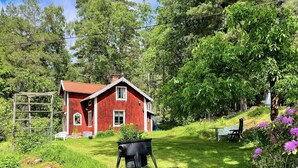 Ett rött hus med en träveranda, omgivet av frodig grönska och en grill.